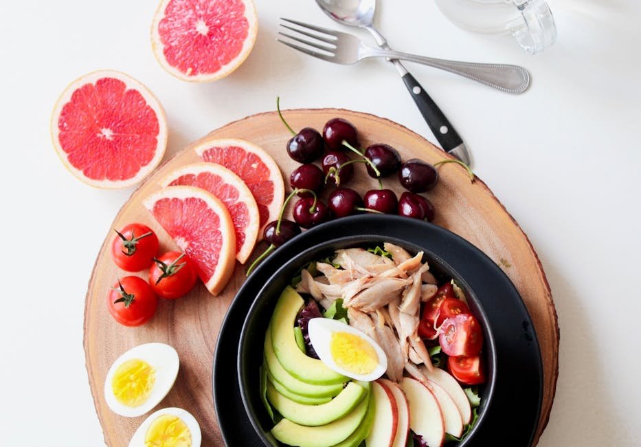 A vibrant breakfast flat lay featuring a mix of fruits, avocado, eggs, tomatoes, and sliced meats on a wooden platter. Balanced diet
